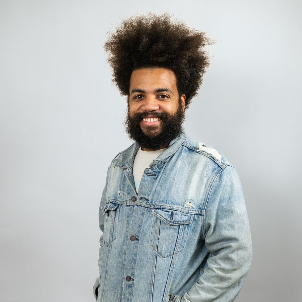 A man with a large afro and beard smiles at the camera, wearing a light denim jacket over a white shirt against a plain light background.