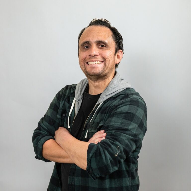 A man with dark hair smiles at the camera with his arms crossed, wearing a black t-shirt, green plaid shirt, and grey hood. Plain light background.