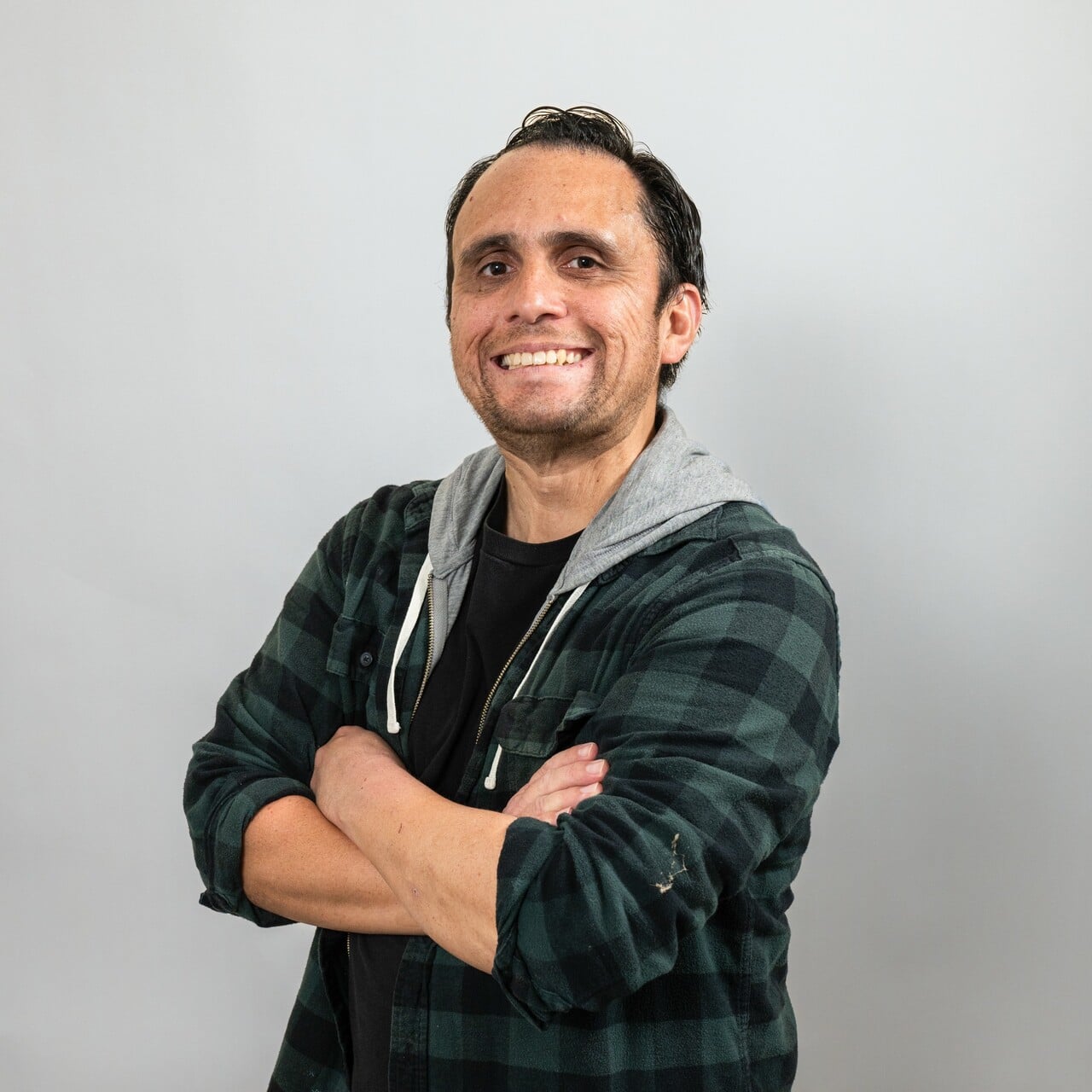 A man with dark hair smiles at the camera with his arms crossed, wearing a black t-shirt, green plaid shirt, and grey hood. Plain light background.