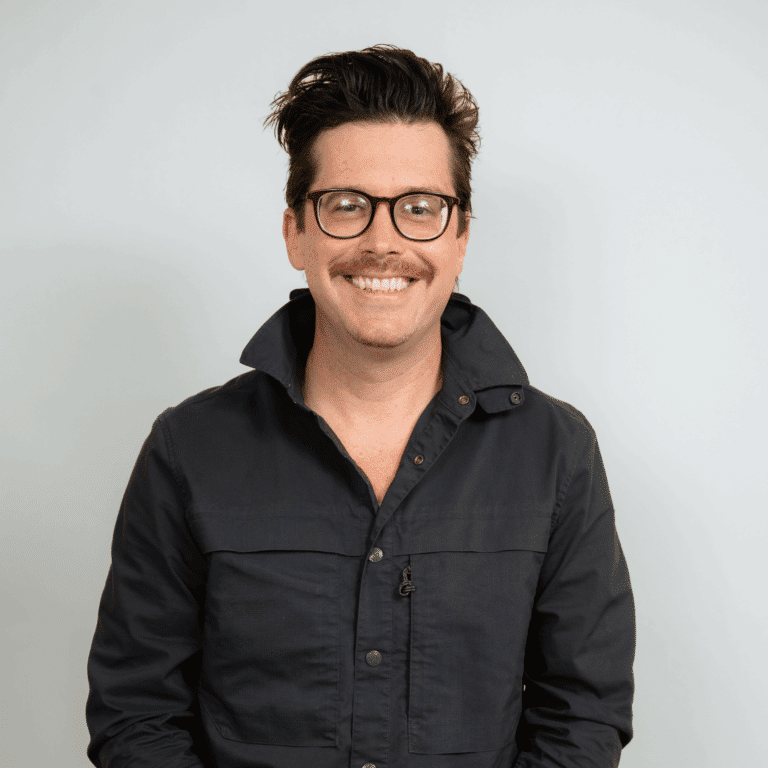 A man with dark hair, glasses, and a mustache is smiling at the camera. He is wearing a dark jacket with the collar slightly raised against a plain light background.