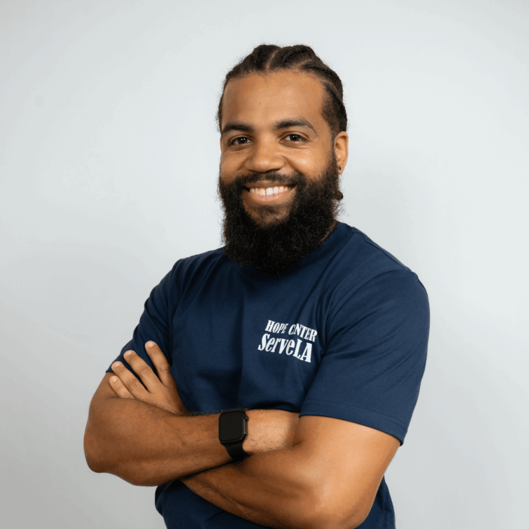 A man with a beard and braided hair, wearing a navy "Hope Center ServeLA" t-shirt and smart watch, stands with arms crossed, smiling at the camera against a plain background.