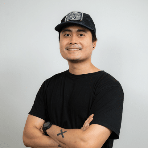 A person wearing a black t-shirt and a black cap, smiling with arms crossed, stands against a plain light background.